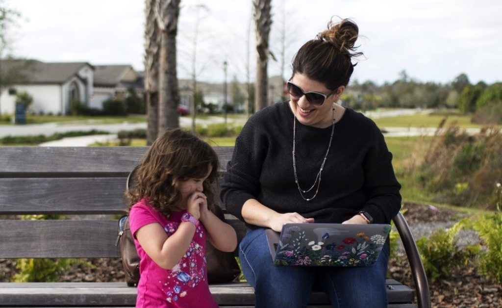 Mother and daughter sitting on a bench, looking at a laptop outdoors.