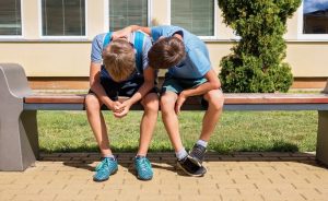 Two boys sitting on a bench with their heads down.