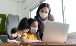 Teacher and student, both wearing masks, using a laptop in a classroom.
