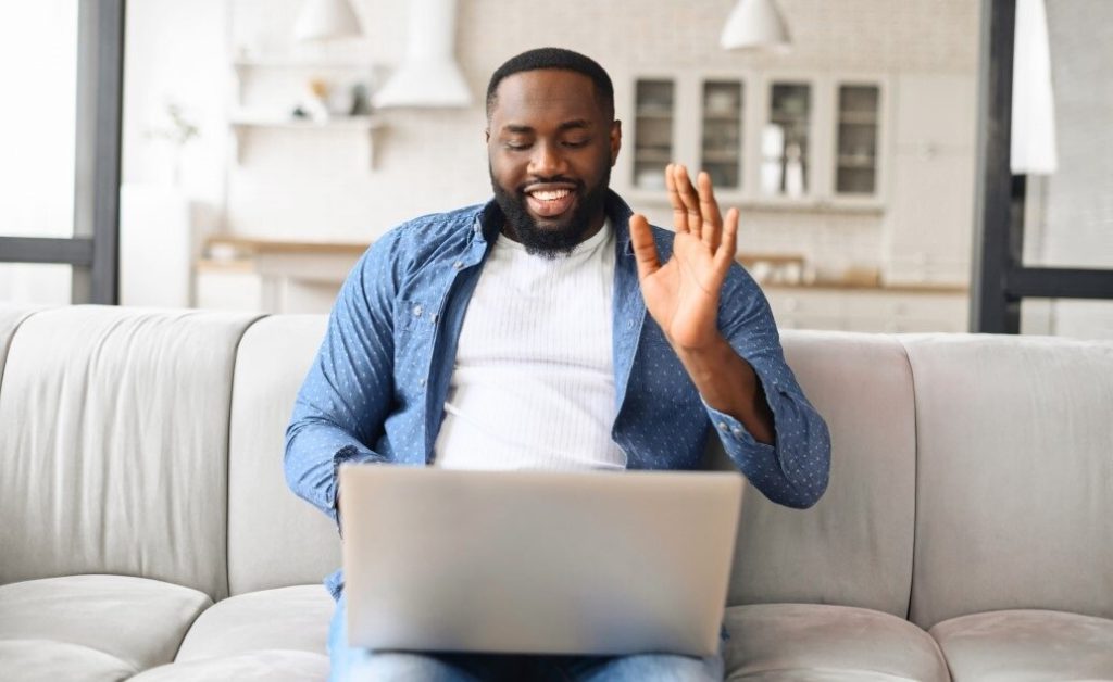 Male on a video call using a laptop, possibly participating in online classes. They are waving with one hand and smiling.