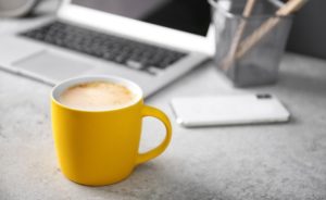 Cup of coffee on desk with laptop in the background.