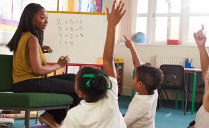 A Black female teacher in a yellow top teaches math at a whiteboard to students with raised hands in a classroom.