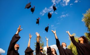 A group of graduates in black gowns throwing their graduation caps in the air against a blue sky.