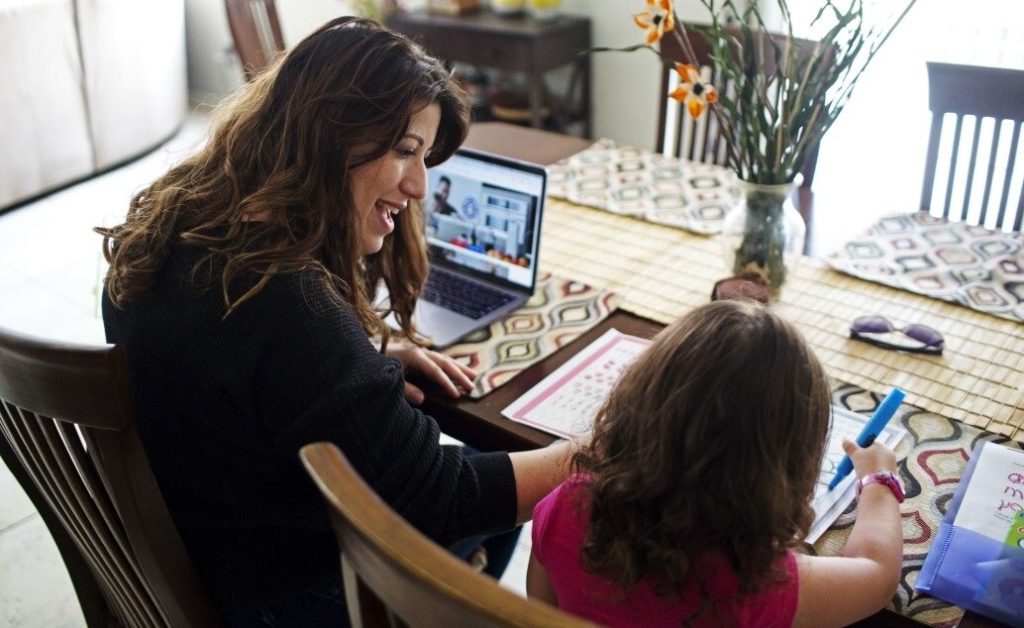 Adult on video call on laptop and child coloring at dining table.