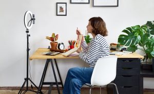 Female teacher recording herself drawing at a desk with a ring light and plants nearby.