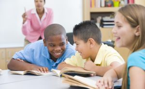A group of students are talking at desks while a female teacher is teaching in background.