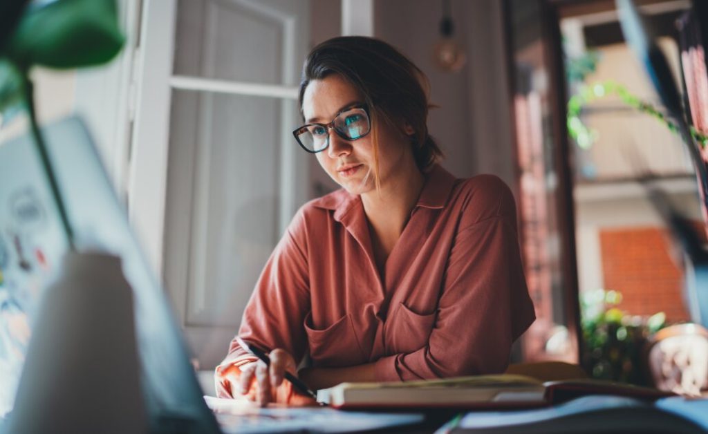 A woman with glasses works at a desk with a laptop and notebook.