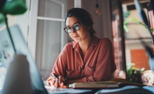 A woman with glasses works at a desk with a laptop and notebook.
