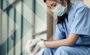 A person wearing medical scrubs, a face mask, and gloves, looking down with a frustrated expression.
