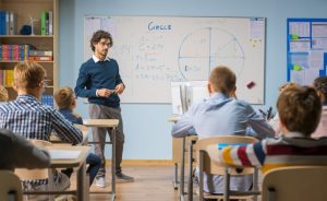 Male teacher standing at the front of a classroom full of students, a whiteboard with mathematical equations is visible.