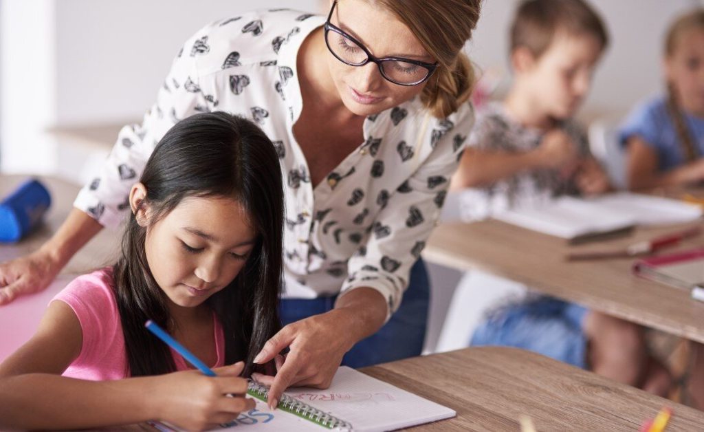 Female teacher helps young girl writing in notebook at desk in classroom with other students.