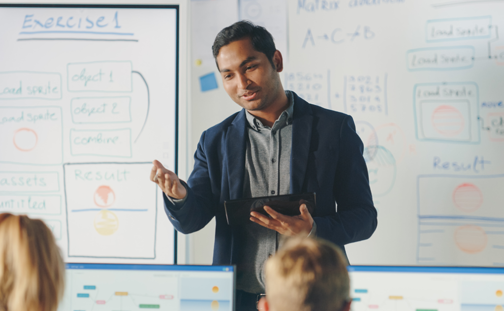 Indian male teacher standing at the front of the classroom, talking to students, with a whiteboard full of text behind him