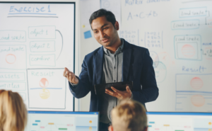 Indian male teacher standing at the front of the classroom, talking to students, with a whiteboard full of text behind him