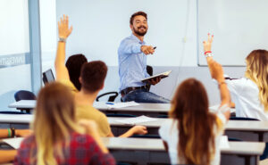 Students raising their hands in a classroom to ask questions while teacher points to one student