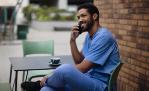 Male nurse in scrubs talking on cell-phone while sitting outside with a coffee.